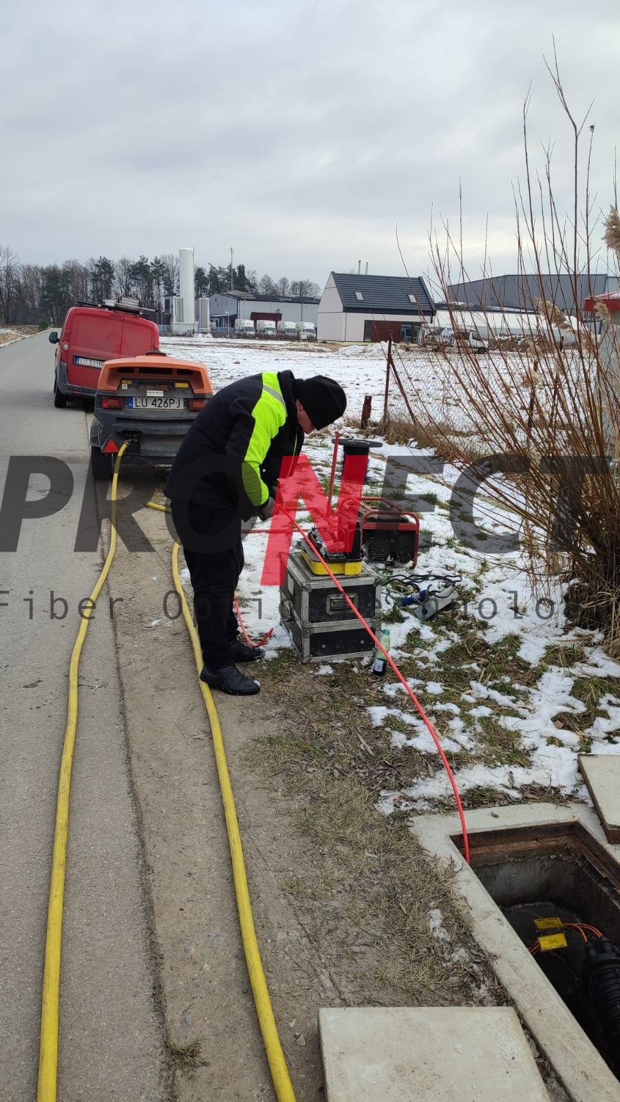 Technik w czarno-żółtej kurtce pochyla się nad skrzynią narzędziową obok otwartej studzienki kanalizacyjnej z widocznymi kablami światłowodowymi, w tle zaparkowany samochód dostawczy z przyczepą...