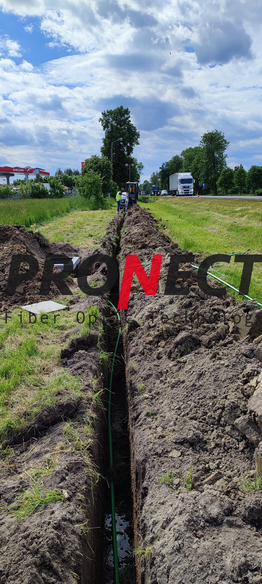 Wykop liniowy w ziemi z zielonym kablem światłowodowym na dnie, w tle pracownicy i maszyna budowlana, po prawej droga z ruchem samochodowym, w oddali stacja benzynowa.