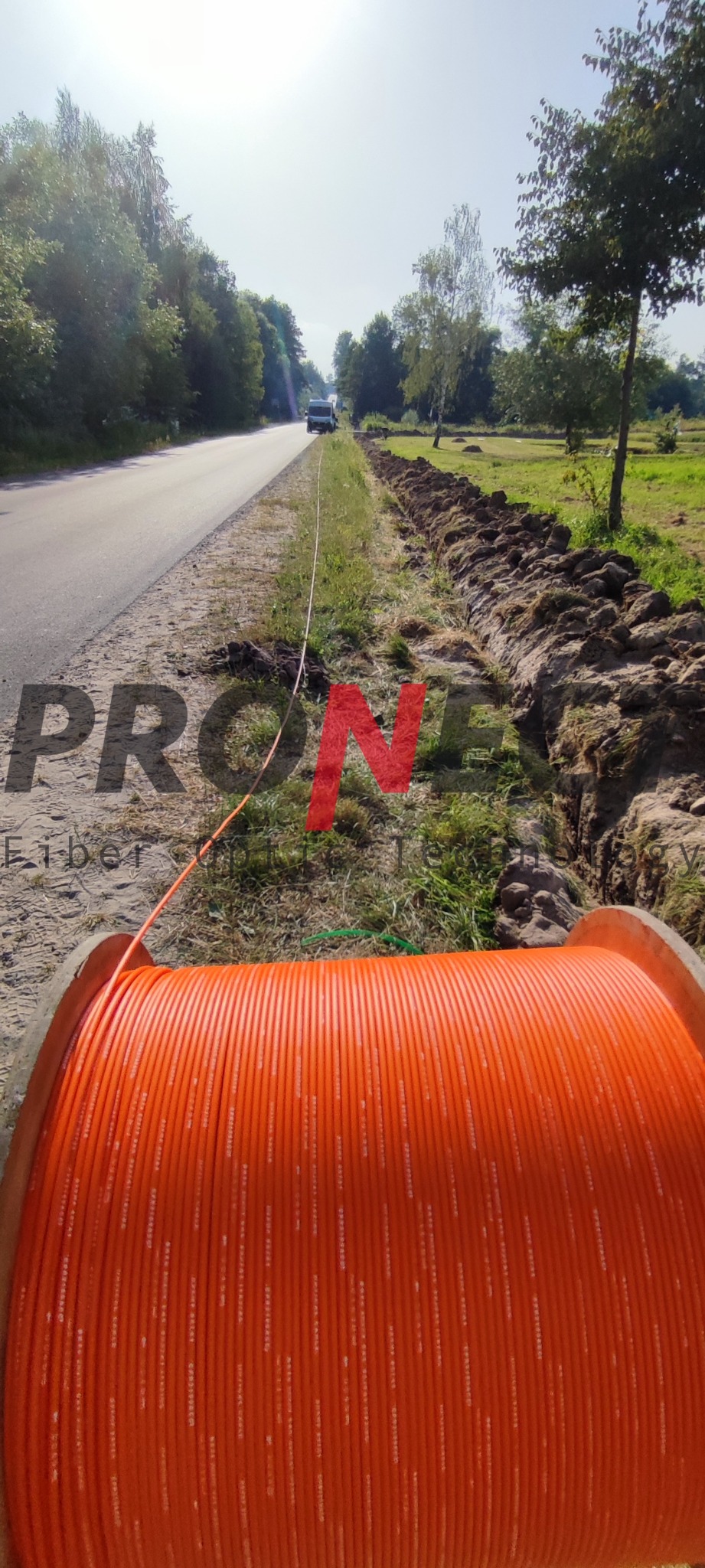 Pomarańczowy bęben z kablem światłowodowym leżący obok wykopu wzdłuż drogi, z widocznym samochodem w oddali, sugerujący prace instalacyjne.