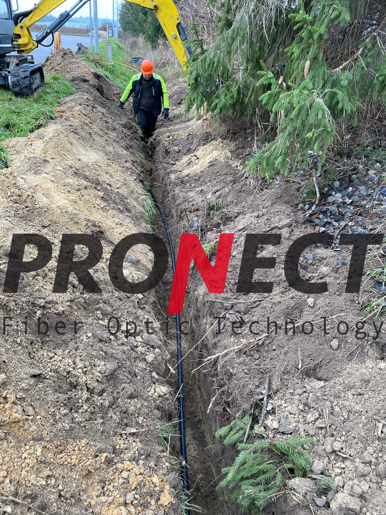 Wykop z ułożonym kablem światłowodowym, pracownik w kasku i kamizelce odblaskowej kontroluje ułożenie kabla, obok stoi mała koparka.