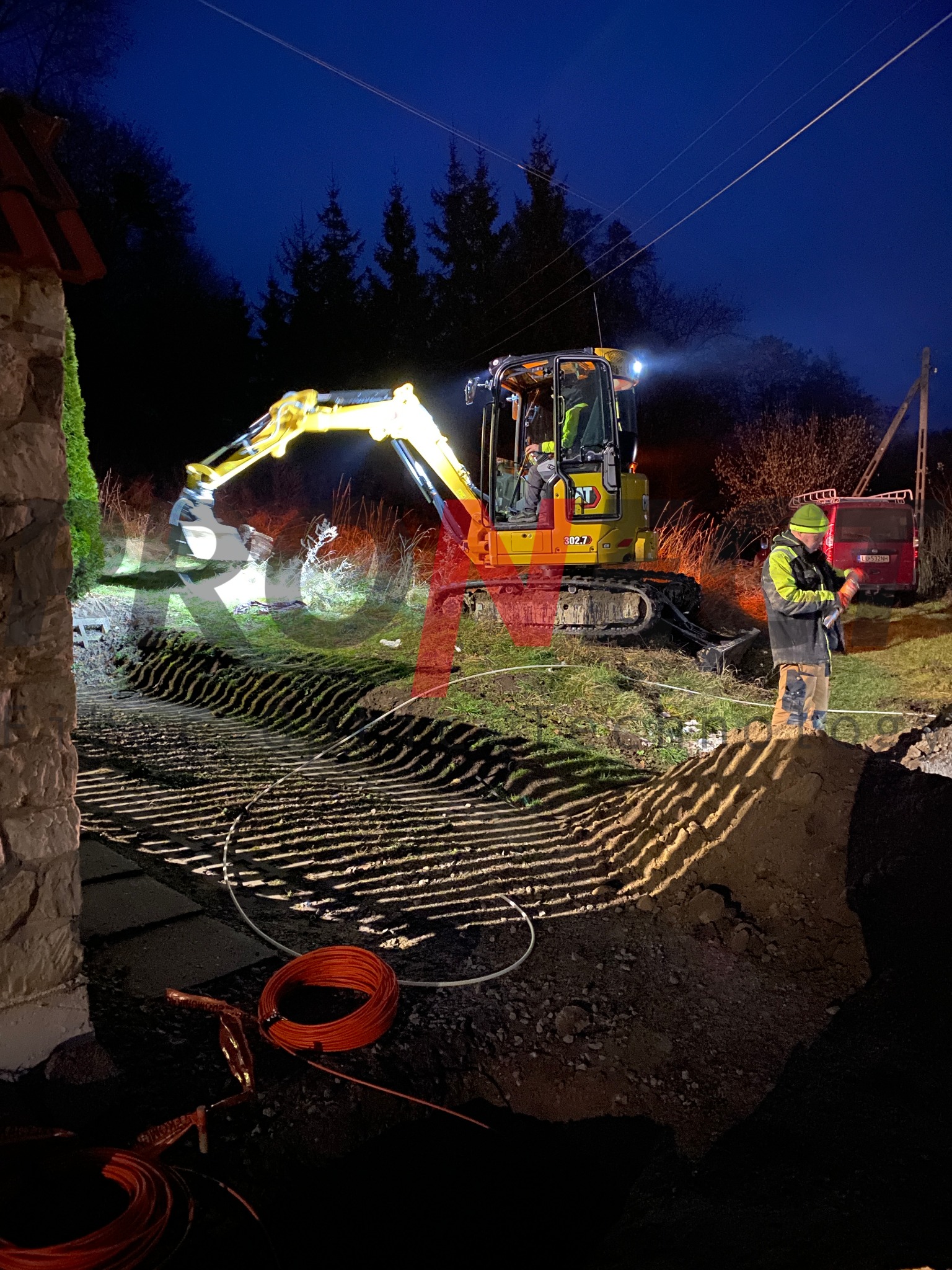 Żółta koparka CAT 302.7 z operatorem w kabinie, oświetlona reflektorami, kopie rów w ziemi. Obok stoi mężczyzna w zielonej czapce i odblaskowej kamizelce. W tle widoczny czerwony samochód i słup...