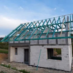 Surowy stan domu z białych bloczków z widoczną konstrukcją dachu z impregnowanego drewna w kolorze turkusowym, na tle zielonych wzgórz i błękitnego nieba.