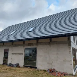 Surowy, niewykończony dom z białego bloczka z zamontowanym, ciemnoszarym dachem z dachówką i dwoma oknami dachowymi, stosy czerwonej dachówki leżące na ziemi.