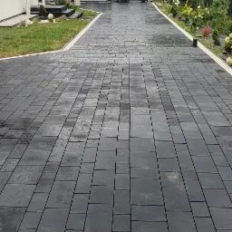 Nowoczesny podjazd z ciemnej kostki brukowej ułożonej we wzór prostokątów, obrzeża z jasnej kostki, w tle dom z białymi ścianami i szary garaż.