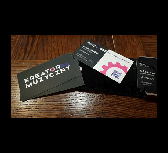 Wizytówki firmowe w czarnym etui na drewnianym stole, z logo Kreator Muzyczny oraz z motywem różowego koła zębatego i kodem QR.