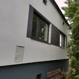 Dwukolorowa elewacja budynku z oknami w ciemnych obramowaniach, skrzynka elektryczna na ścianie, drewniana ławka i szpadel przy ścianie.