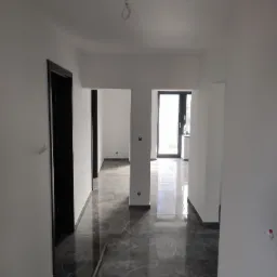 Nowoczesny korytarz z szarą marmurową podłogą, świeżo pomalowanymi białymi ścianami i czarnymi futrynami drzwi, widok na jasne pomieszczenie z oknem.