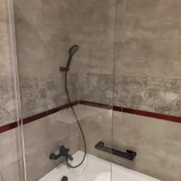 Wykończona łazienka z wanną obudowaną płytkami, czarna armatura i szklany parawan nawannowy z półką na kosmetyki.