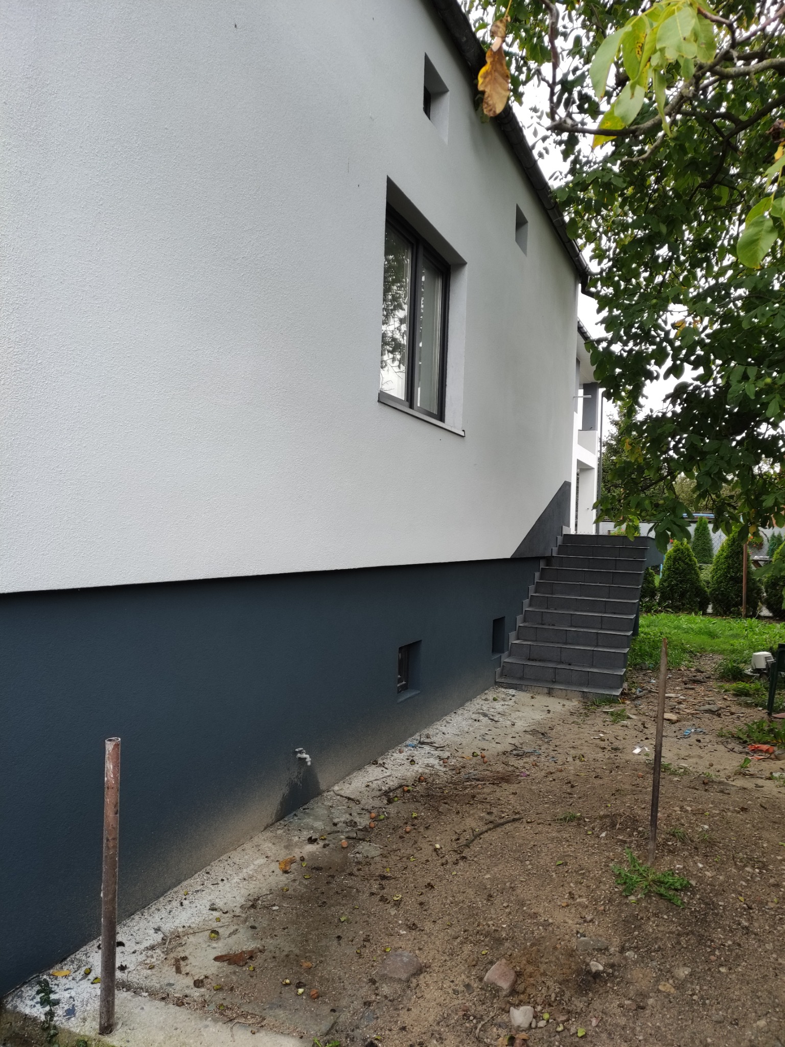 Wykończona elewacja budynku z dwukolorowym tynkiem, widoczne schody wejściowe z szarej kostki i fragment ogrodu z drzewem w tle.