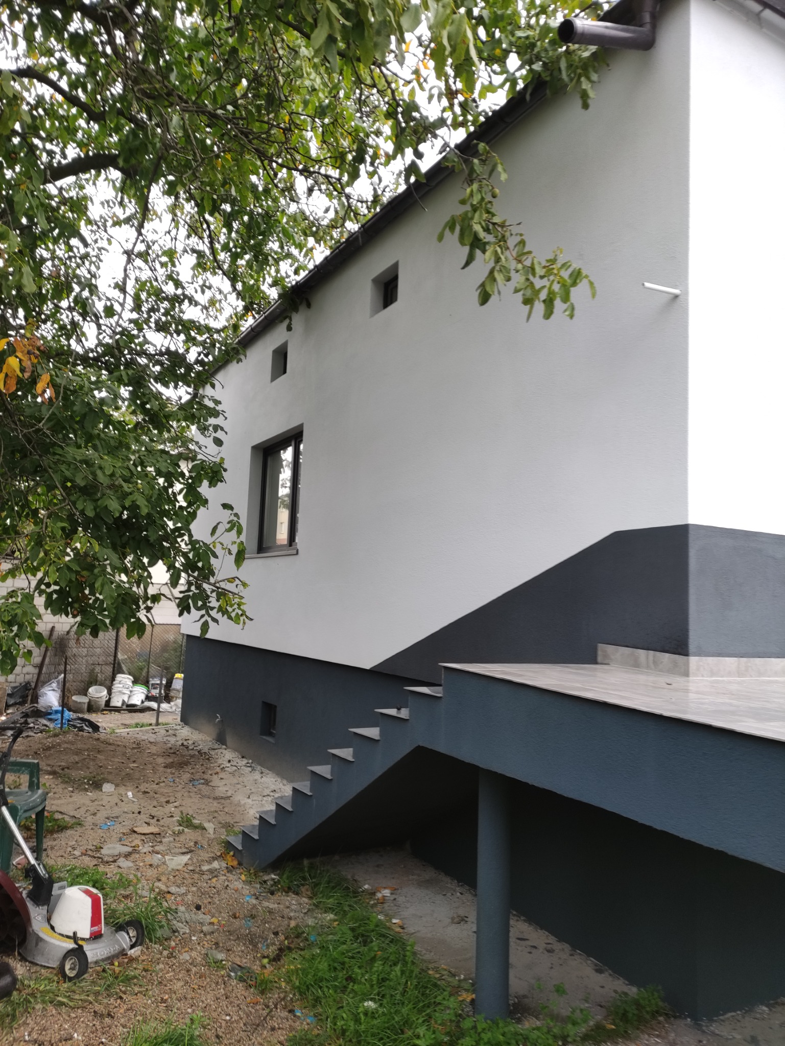Ocieplony budynek z widocznym fragmentem elewacji pomalowanym na szaro, schody prowadzące do wejścia, trawnik z kosiarką i drzewo z zielonymi liśćmi.