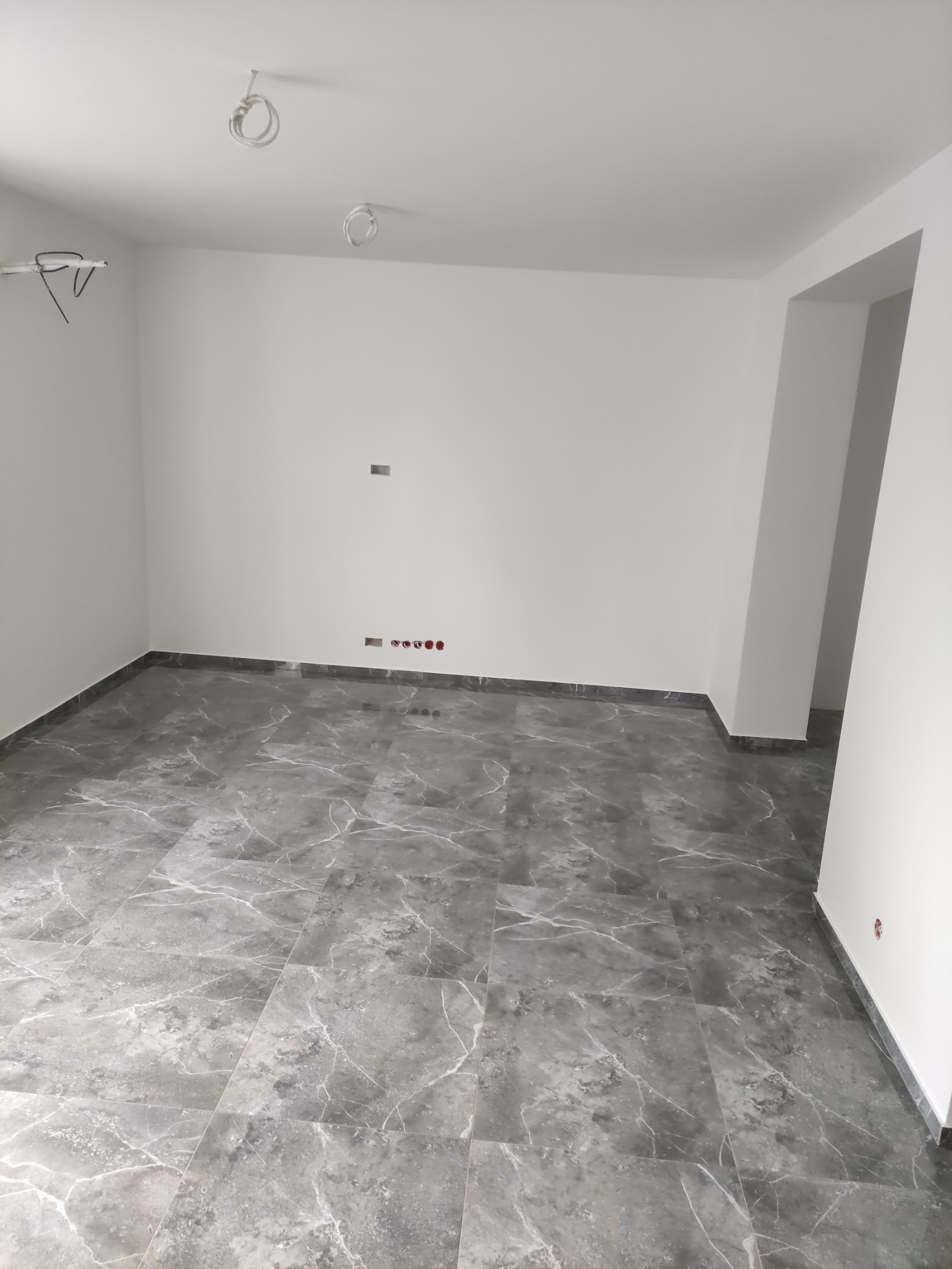 Pokój z szarą marmurową podłogą w trakcie wykańczania, widoczne kable wystające z sufitu i gniazdka elektryczne na ścianie.
