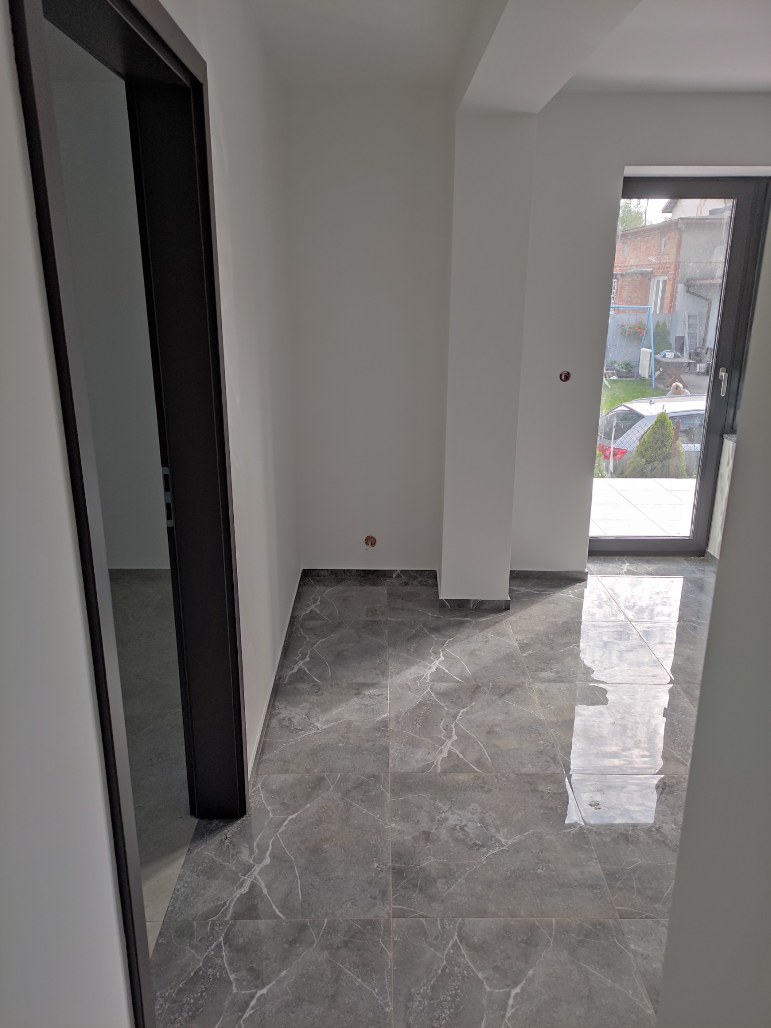 Wykończone wnętrze: szare płytki podłogowe z marmurkowym wzorem, białe ściany z widocznymi puszkami instalacyjnymi, czarna rama drzwiowa i okienna z widokiem na ogród.