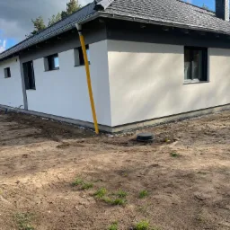 Nowoczesny dom jednorodzinny z białą elewacją i grafitowymi elementami wykończeniowymi, widoczne rury spustowe w kolorze żółtym, teren wokół domu w trakcie zagospodarowywania.