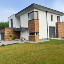 Nowoczesny dom z białymi ścianami i elementami z cegły klinkierowej, szara stolarka okienna i drzwiowa, minimalistyczny ogród z trawnikiem i młodym drzewkiem.