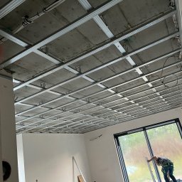 KB KOMPLEKSOWA REALIZACJA BUDÓW KAMIL BARTKOWIAK - Montaż metalowej konstrukcji sufitu podwieszanego z widocznymi przewodami, w tle pracownik przy oknie.