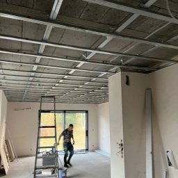 KB KOMPLEKSOWA REALIZACJA BUDÓW KAMIL BARTKOWIAK - Montaż metalowego rusztu sufitowego podwieszanego, widoczna drabina i sylwetka osoby w tle, surowy stan wykończenia wnętrza.