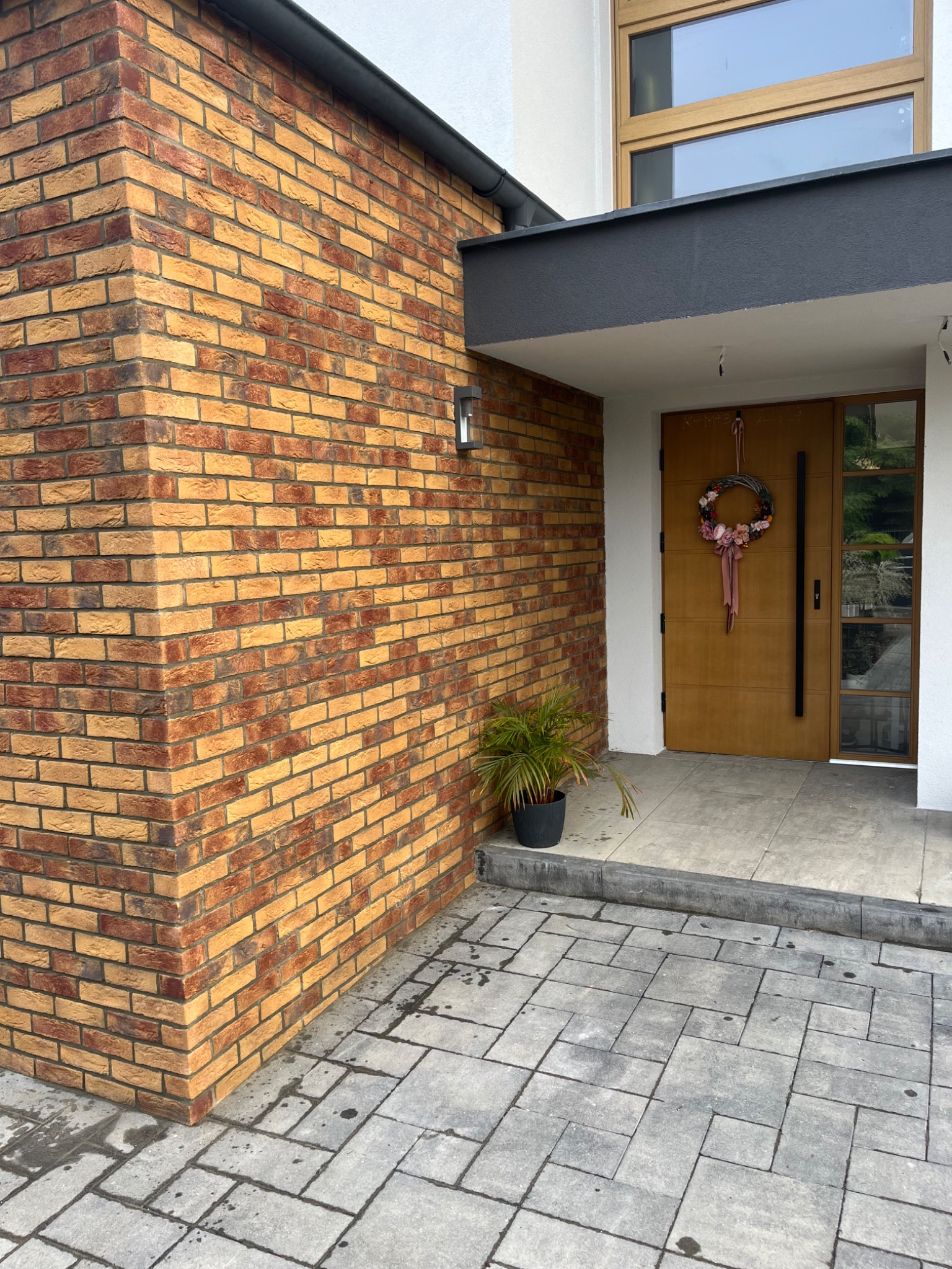 Wejście do domu z elewacją z cegły klinkierowej w odcieniach żółci i czerwieni, nowoczesne drzwi z wieńcem i rośliną w doniczce, bruk na podjeździe.