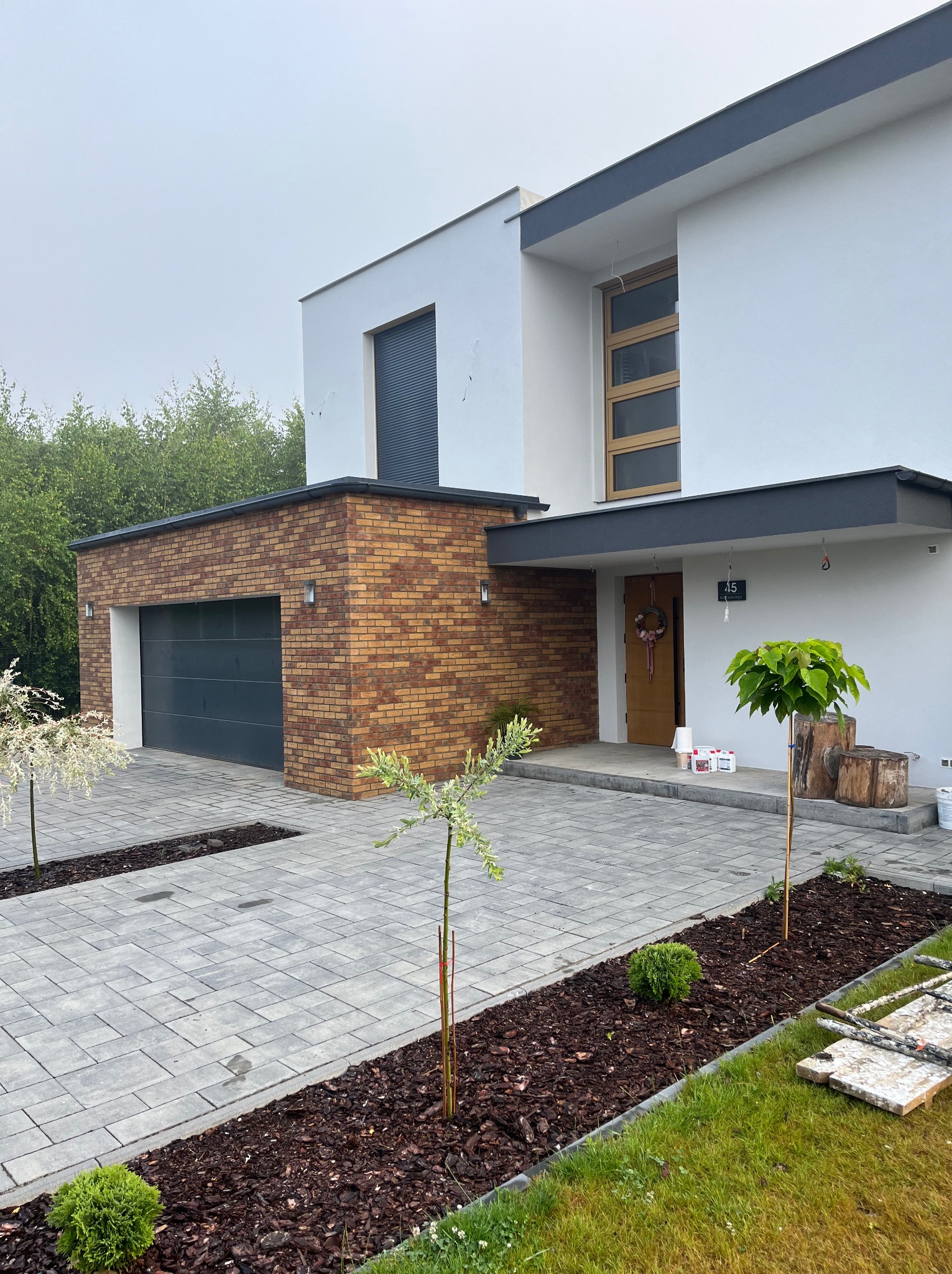 Nowoczesny dom murowany z ceglanym garażem i szarą bramą, wykończony białą elewacją i szarymi akcentami, z brukowanym podjazdem i młodymi drzewkami.