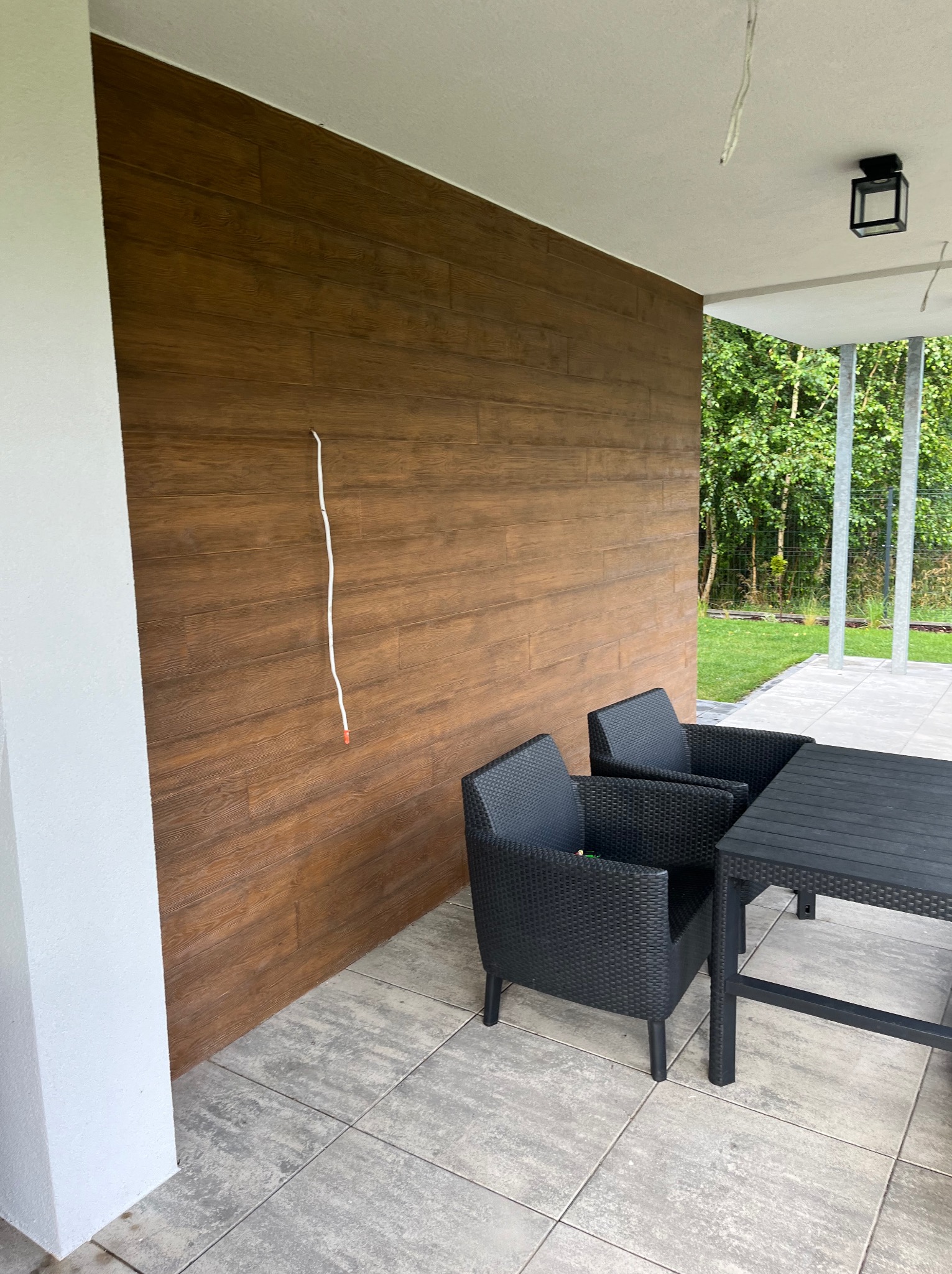 Zadaszony taras z drewnopodobną okładziną elewacyjną, wyposażony w czarne meble ogrodowe i szarą kostkę brukową; widoczny wystający kabel ze ściany.