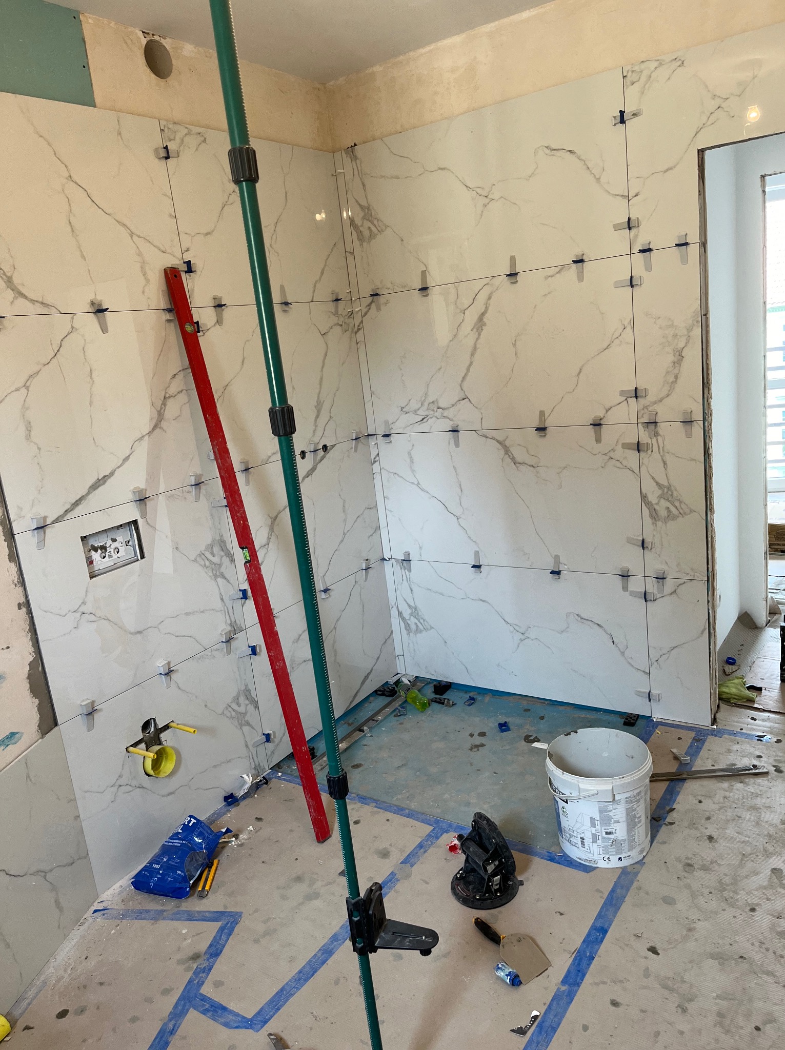 Ściana łazienki w trakcie wykańczania z białymi płytkami imitującymi marmur, poziomica oparta o ścianę, widoczne narzędzia glazurnicze, krzyżyki dystansowe i system poziomowania płytek na ścianach...