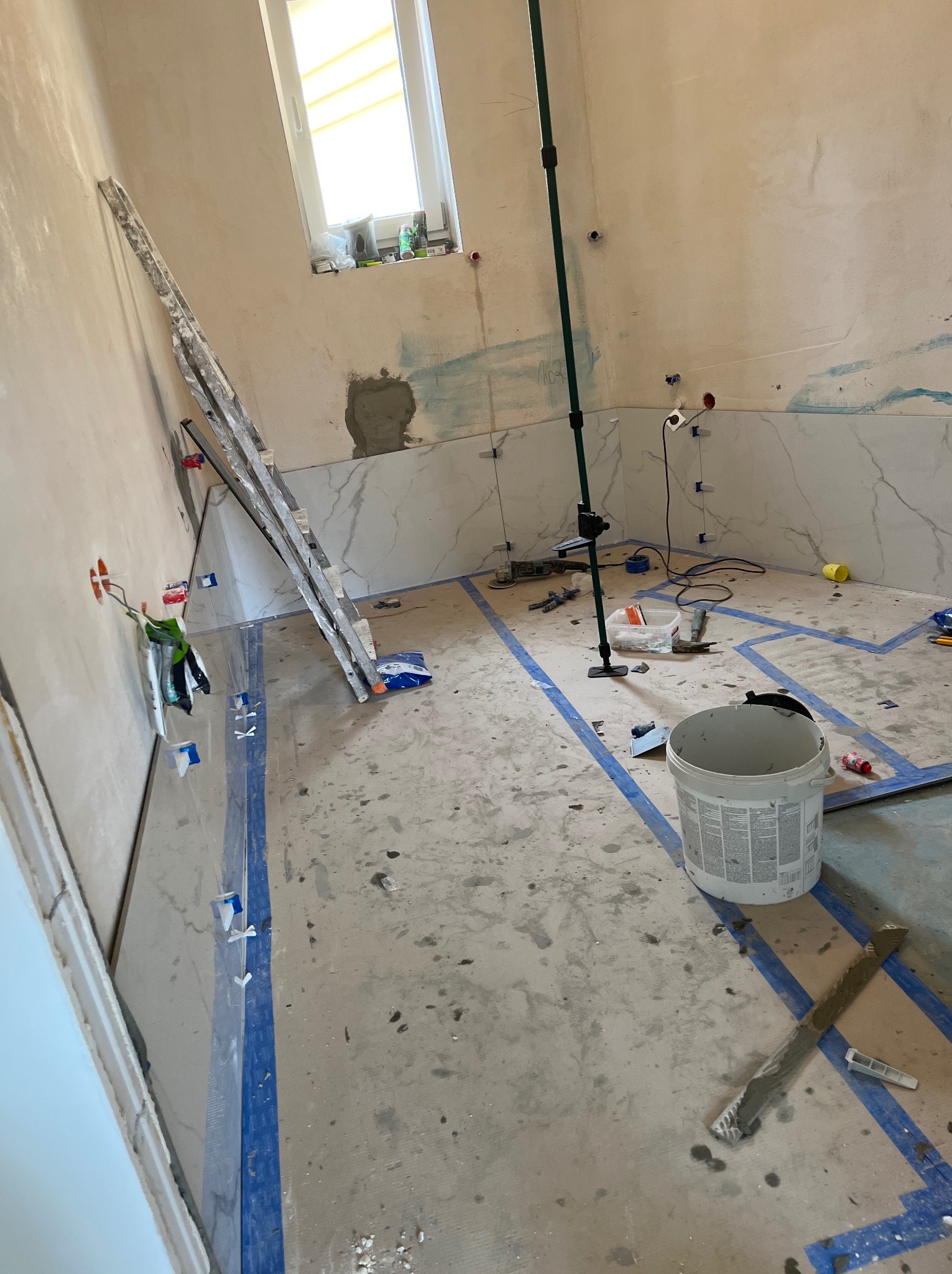 Łazienka w trakcie układania marmurowych płytek, widoczne narzędzia, poziomica, wiadro z klejem i taśma malarska wyznaczająca obszar roboczy.