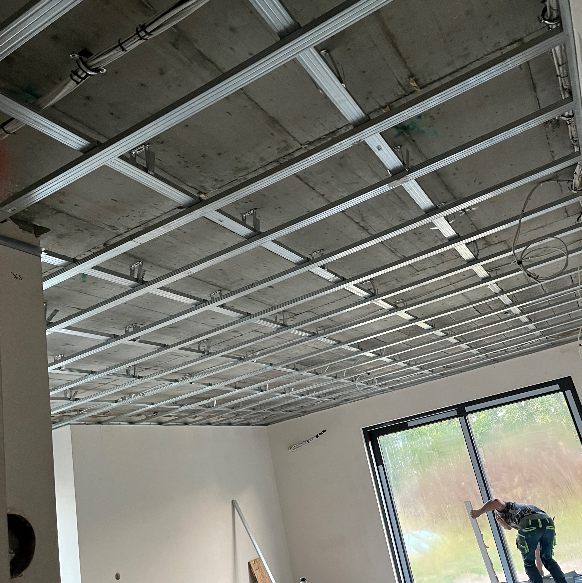 Montaż metalowej konstrukcji sufitu podwieszanego z widocznymi przewodami, w tle pracownik przy oknie.