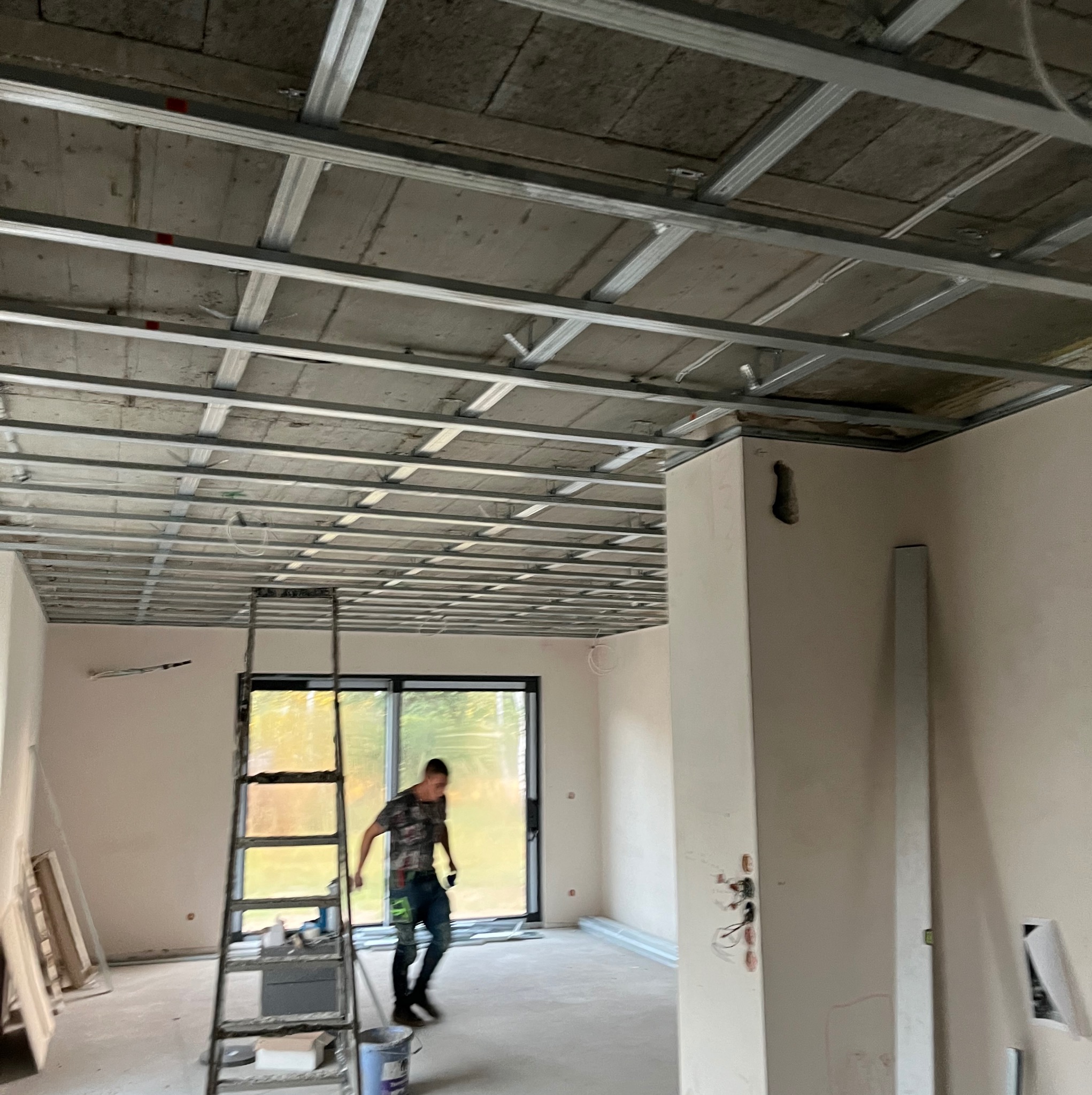 Montaż metalowego rusztu sufitowego podwieszanego, widoczna drabina i sylwetka osoby w tle, surowy stan wykończenia wnętrza.