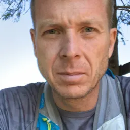 Mężczyzna z krótkimi blond włosami, lekko spocony, w sportowej koszulce i plecaku biegowym, robiący selfie pod drzewem.