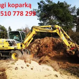 Żółta koparka marki Baustra pracuje na piaszczystym terenie, w tle widoczne drzewa i niebieskie niebo, u góry czerwony napis z numerem telefonu.