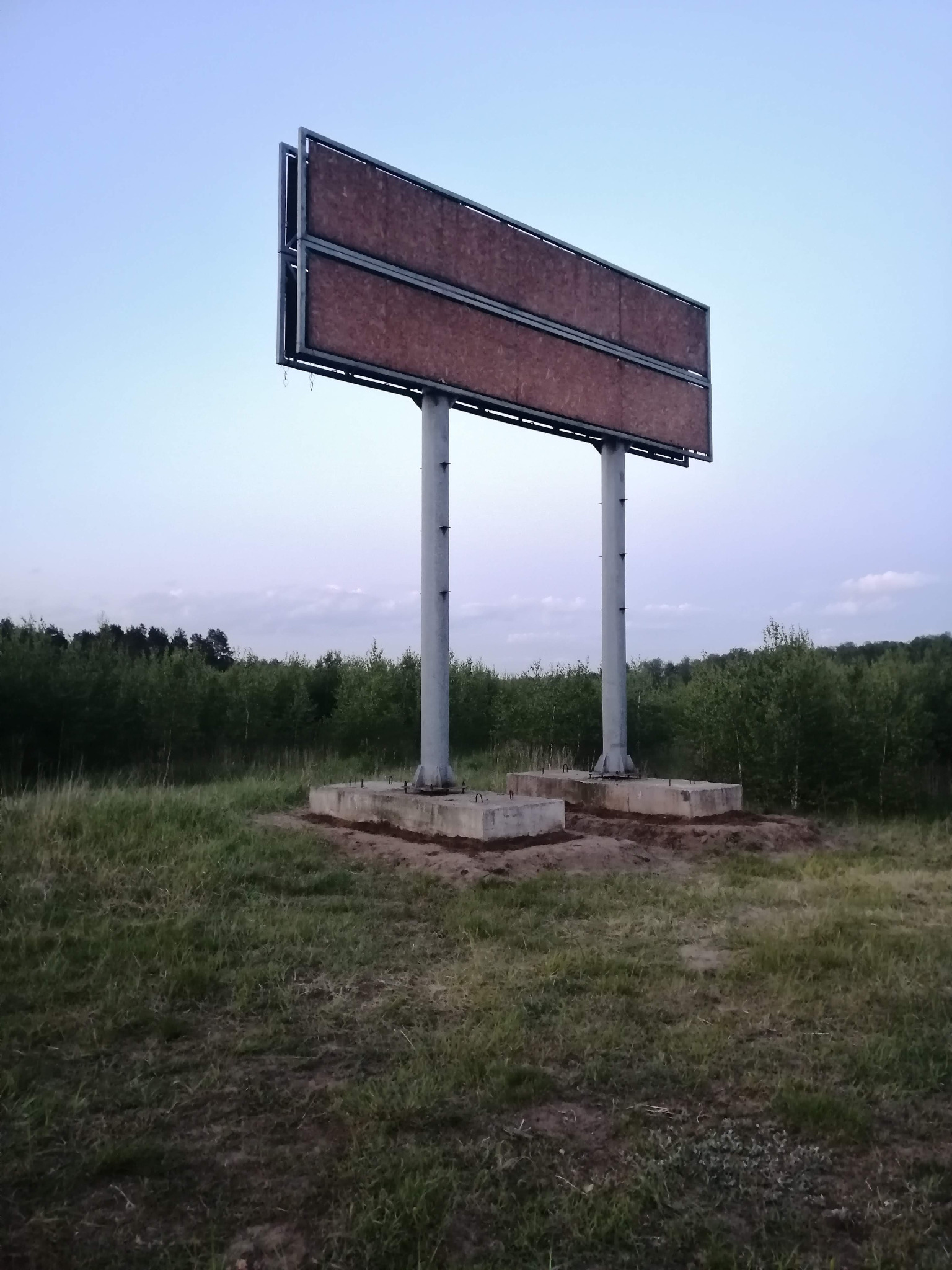 Konstrukcja dwupiętrowego billboardu z brązowym wypełnieniem, na betonowych fundamentach, na tle łąki i lasu, w pochmurny dzień.