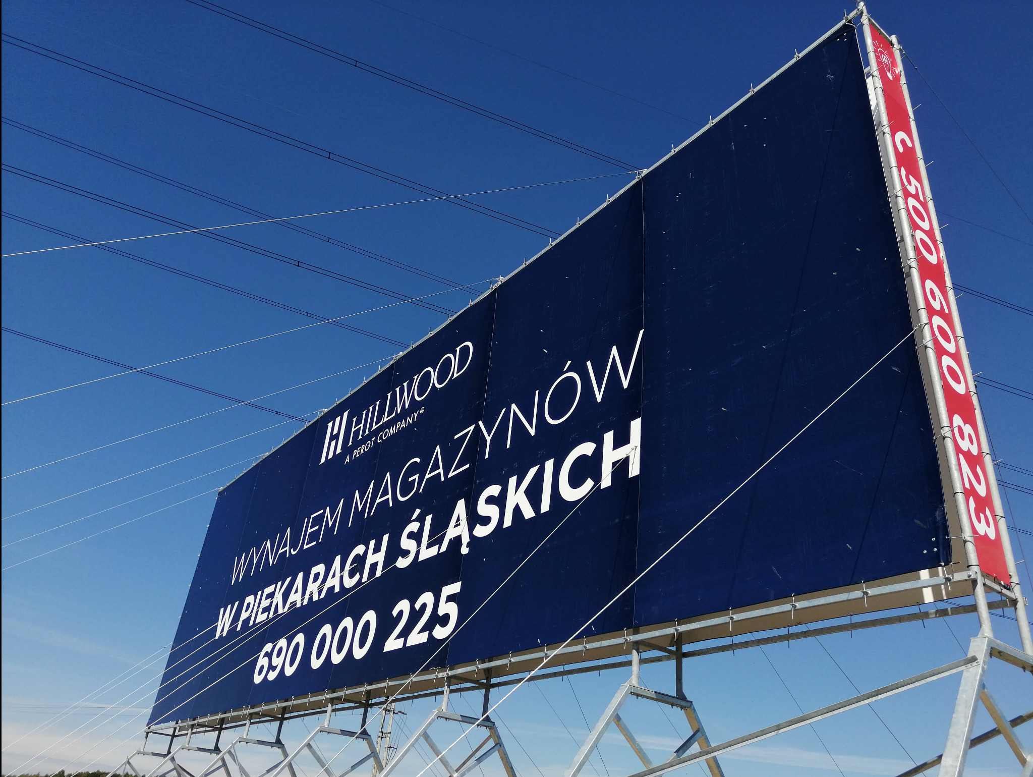 Billboard reklamujący wynajem magazynów Hillwood w Piekarach Śląskich, widoczny numer telefonu, konstrukcja stalowa, błękitne niebo w tle, widok z dołu.