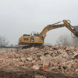 Żółta koparka Komatsu PC 360 NLC rozbiera ceglany budynek, gruzowisko na pierwszym planie, szare niebo w tle.