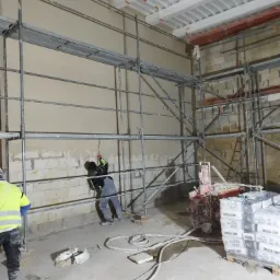 Praca na rusztowaniu: robotnik w trakcie nakładania tynku na ścianę z cegły, widoczna maszyna do tynkowania i paleta worków z zaprawą.