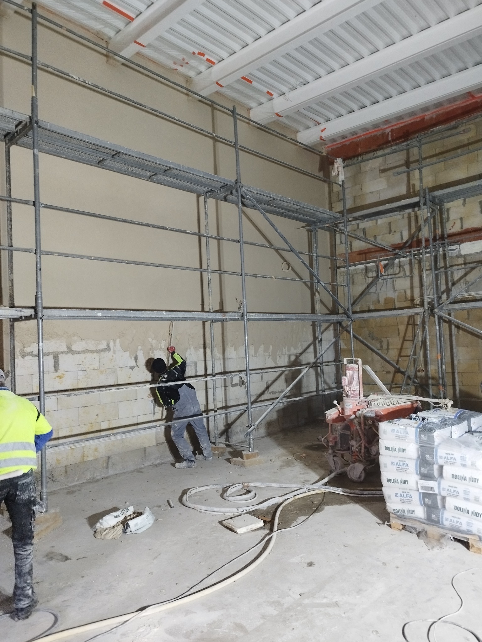 Praca na rusztowaniu: robotnik w trakcie nakładania tynku na ścianę z cegły, widoczna maszyna do tynkowania i paleta worków z zaprawą.