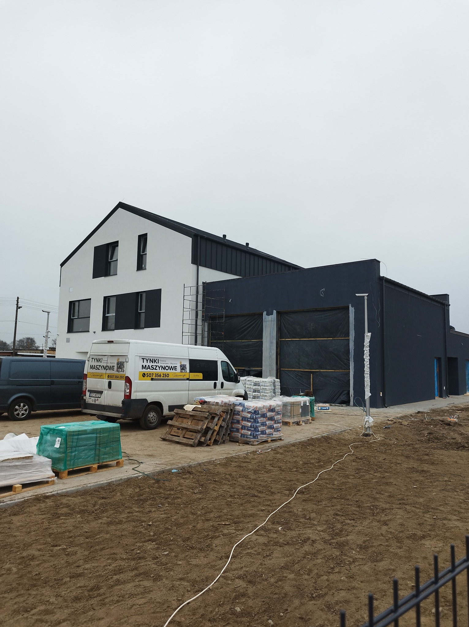 Nowoczesny budynek w trakcie prac wykończeniowych, z zaparkowanym vanem z reklamą firmy, paletami z materiałami budowlanymi i przygotowanym terenem.