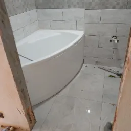 Narożna wanna akrylowa w trakcie montażu w łazience, widoczne szare płytki na ścianach i podłodze, narzędzia glazurnicze i niedokończone obramowanie drzwi.