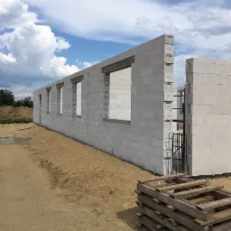 Wznoszenie ścian parteru z betonu komórkowego, widoczne otwory okienne i paleta drewniana na piaszczystym podłożu, błękitne niebo z obłokami w tle.