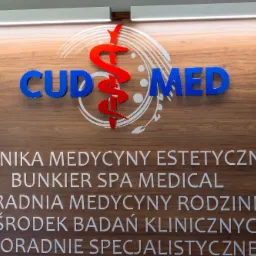 Tablica informacyjna kliniki medycyny estetycznej CUD MED z logo w kolorach czerwonym i niebieskim na drewnianym tle.