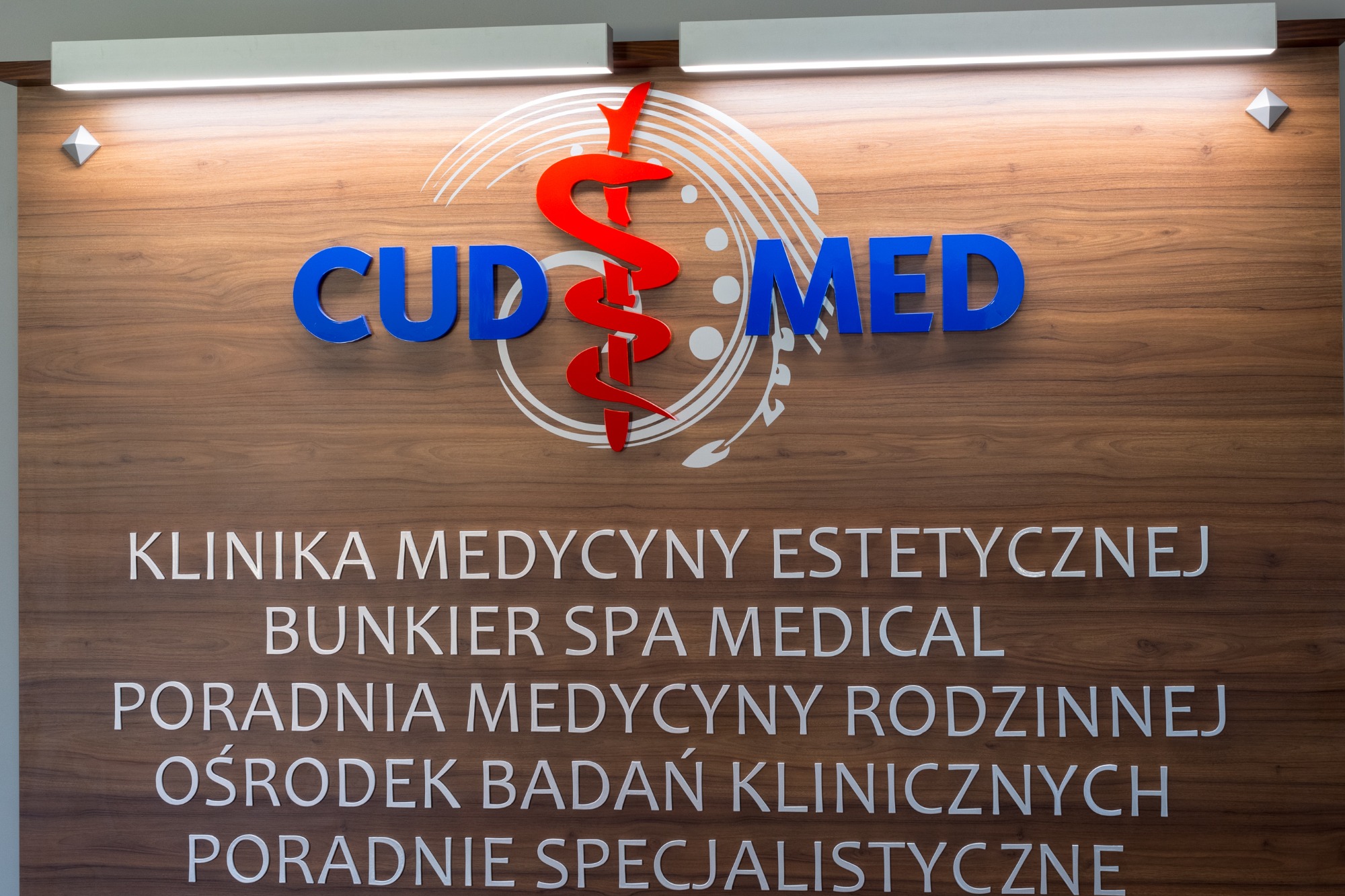 Tablica informacyjna kliniki medycyny estetycznej CUD MED z logo w kolorach czerwonym i niebieskim na drewnianym tle.