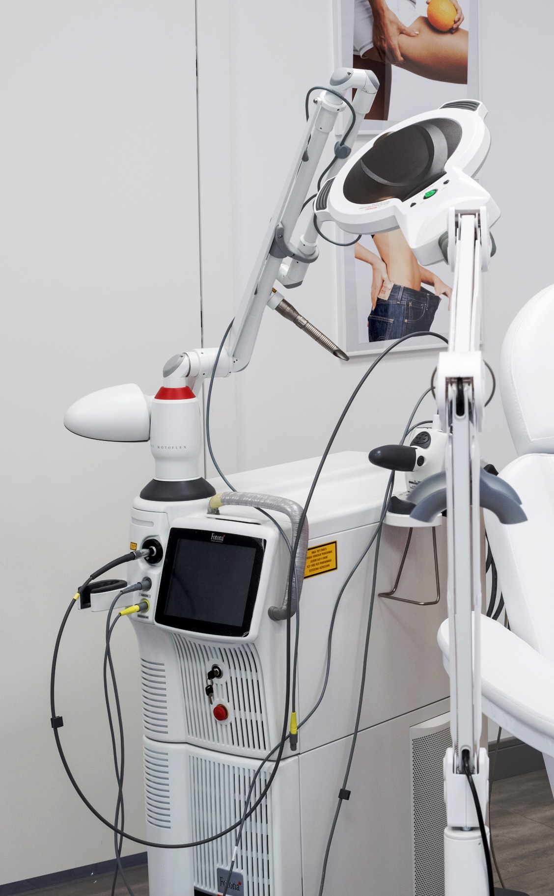 Biały laser kosmetyczny Fotona Rotoflex z ruchomym ramieniem i panelem dotykowym, obok fotel kosmetyczny, w tle plakat z efektami zabiegów.