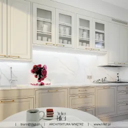 Elegancka kuchnia w odcieniach bieli i szarości z marmurowym blatem, złotymi uchwytami i przeszklonymi szafkami wiszącymi, uzupełniona bukietem róż i filiżanką kawy na okrągłym stole.