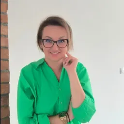 Uśmiechnięta kobieta w okularach i zielonej koszuli, z zegarkiem na ręce, pozuje w jasnym wnętrzu z fragmentem ściany z cegły.