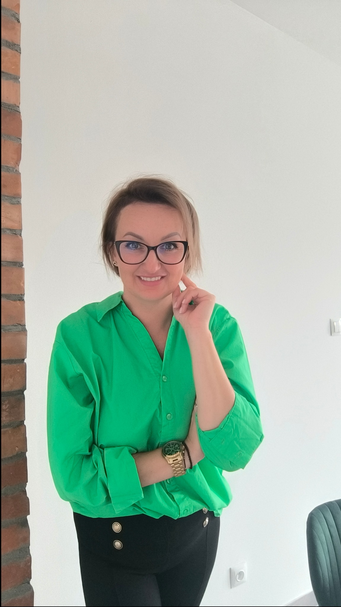 Uśmiechnięta kobieta w okularach i zielonej koszuli, z zegarkiem na ręce, pozuje w jasnym wnętrzu z fragmentem ściany z cegły.