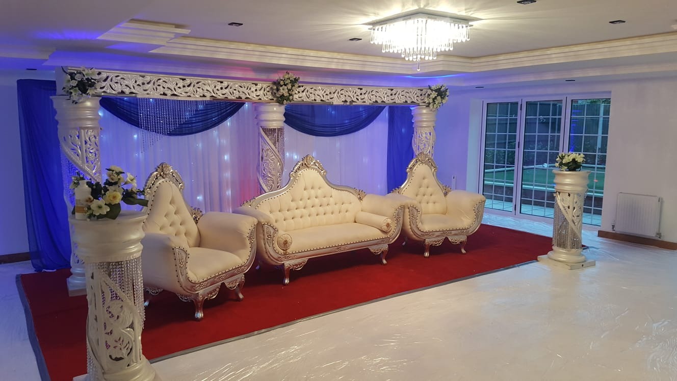 Uroczyste ustawienie: kremowe, pikowane sofy i fotele z dekoracyjnymi srebrnymi obramowaniami, umieszczone na czerwonym dywanie pod ozdobnym baldachimem z białymi kolumnami, kwiatami i niebieskimi...