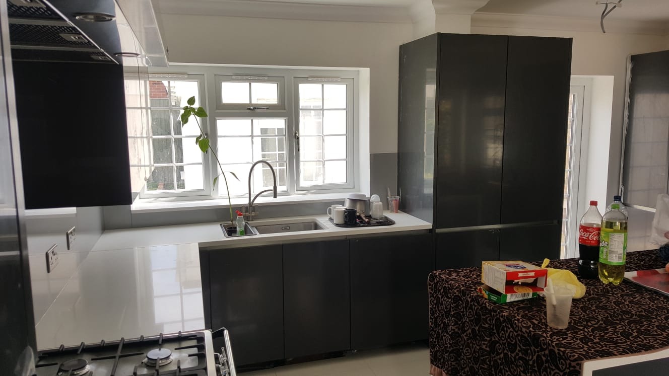 Nowoczesna kuchnia z szarymi szafkami, białym blatem i oknem z widokiem na ceglany budynek. Na blacie zlewozmywak z rosnącą rośliną w doniczce, naczynia i artykuły spożywcze na stole w tle.