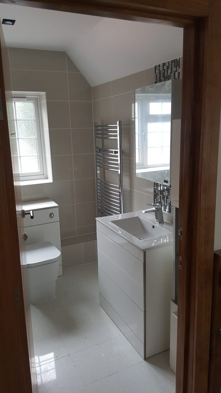 Nowoczesna łazienka z beżowymi płytkami, białą umywalką z szafką, grzejnikiem drabinkowym i lustrem z mozaiką, widok zza drewnianej framugi drzwi.