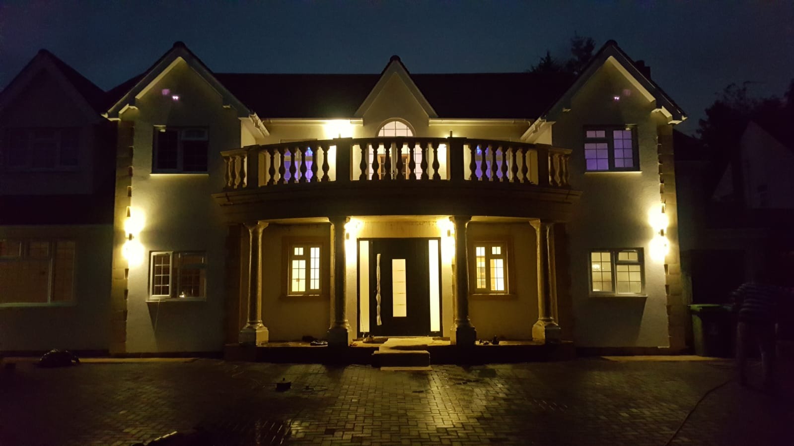 Dwupiętrowy dom z balkonem oświetlony zewnętrznymi lampami w nocy, widoczny bruk na podjeździe.