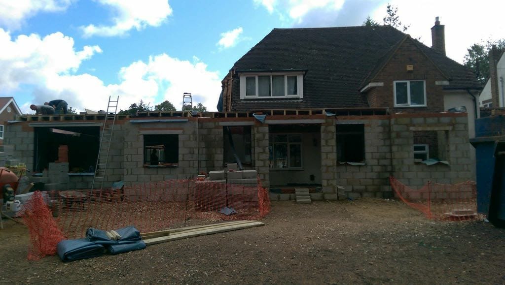 Rozbudowa domu jednorodzinnego w trakcie budowy, widoczne ściany z bloczków betonowych, drewniana konstrukcja dachu, drabina oparta o budynek, robotnicy na dachu, zabezpieczenia budowy.