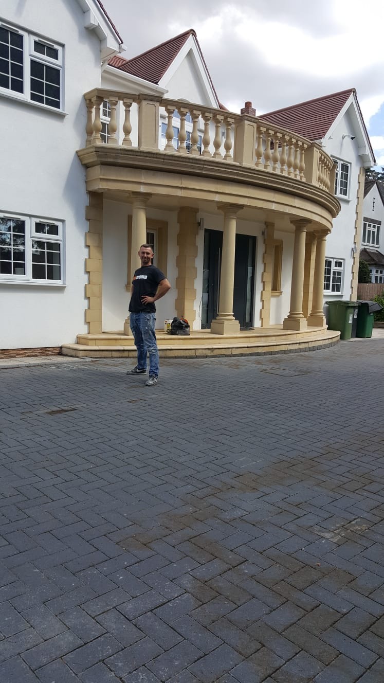 Mężczyzna pozuje na tle odnowionej fasady domu z kolumnami i balkonem z balustradą, widoczny sprzęt budowlany i kostka brukowa.