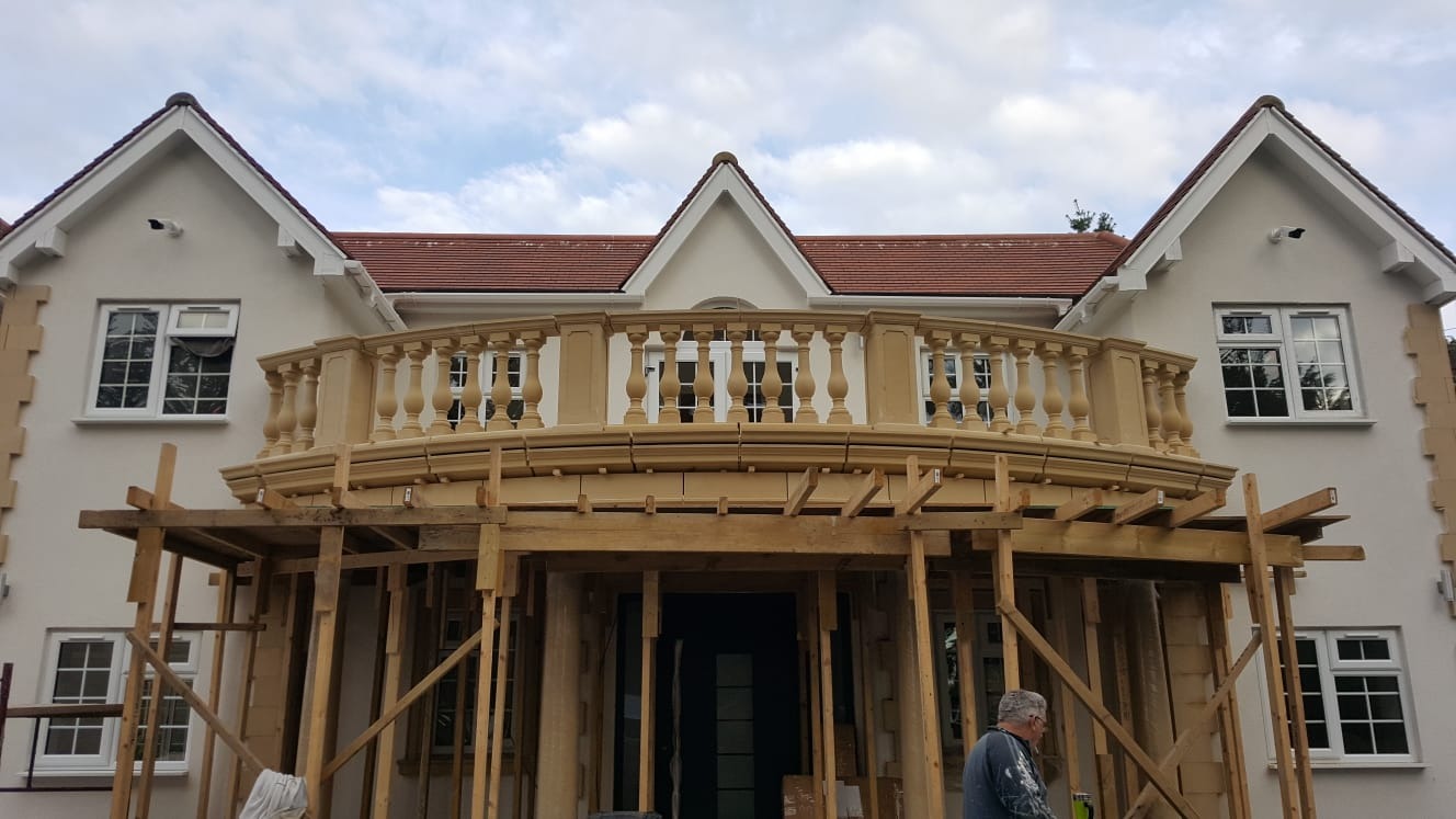Elewacja domu z beżowym balkonem z balustradą w stylu klasycznym, w trakcie montażu rusztowania, widoczny pracownik w dolnym rogu.